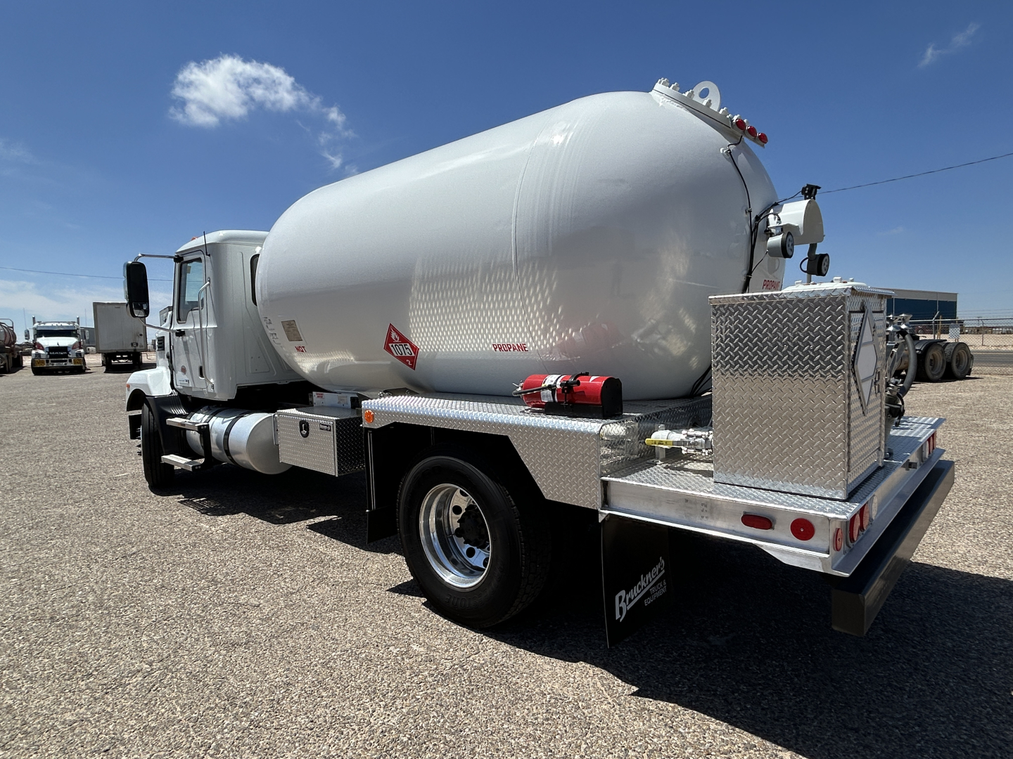Tank truck chassis with cylindrical rear tank and rear plumbing, Mack MD7 vehicle in background showing fuel or liquid storage system.