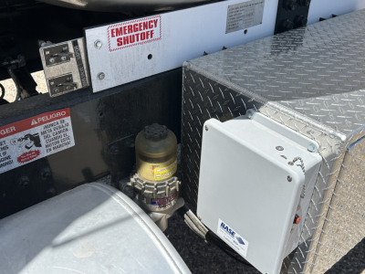 Mack MD7 truck engine bay area showing emergency shutoff lever, fuel/air lines, and a rectangular control box with a base label. Diamond plate toolbox and hydraulic components visible.