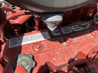 Engine bay of a new 2026 Mack MD7 showing the red engine cover with label and oil cap. Visible text includes 'B6.7' and various warning labels near the top.