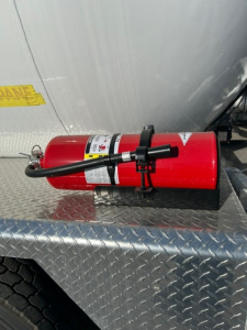 Fire extinguisher mounted on diamond plate; red cylinder with labels and handle, hose attached, on truck side near cabin