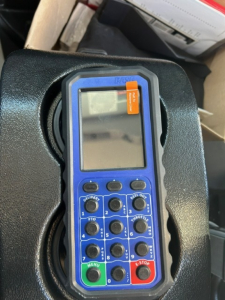 Blue handheld remote control device with numerous black buttons and small LCD screen; orange label at top-right corner; device sits in a vehicle compartment.