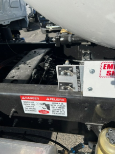 Truck chassis area with warning label and control switches visible under a tank, showing danger sticker and a dual switch panel.