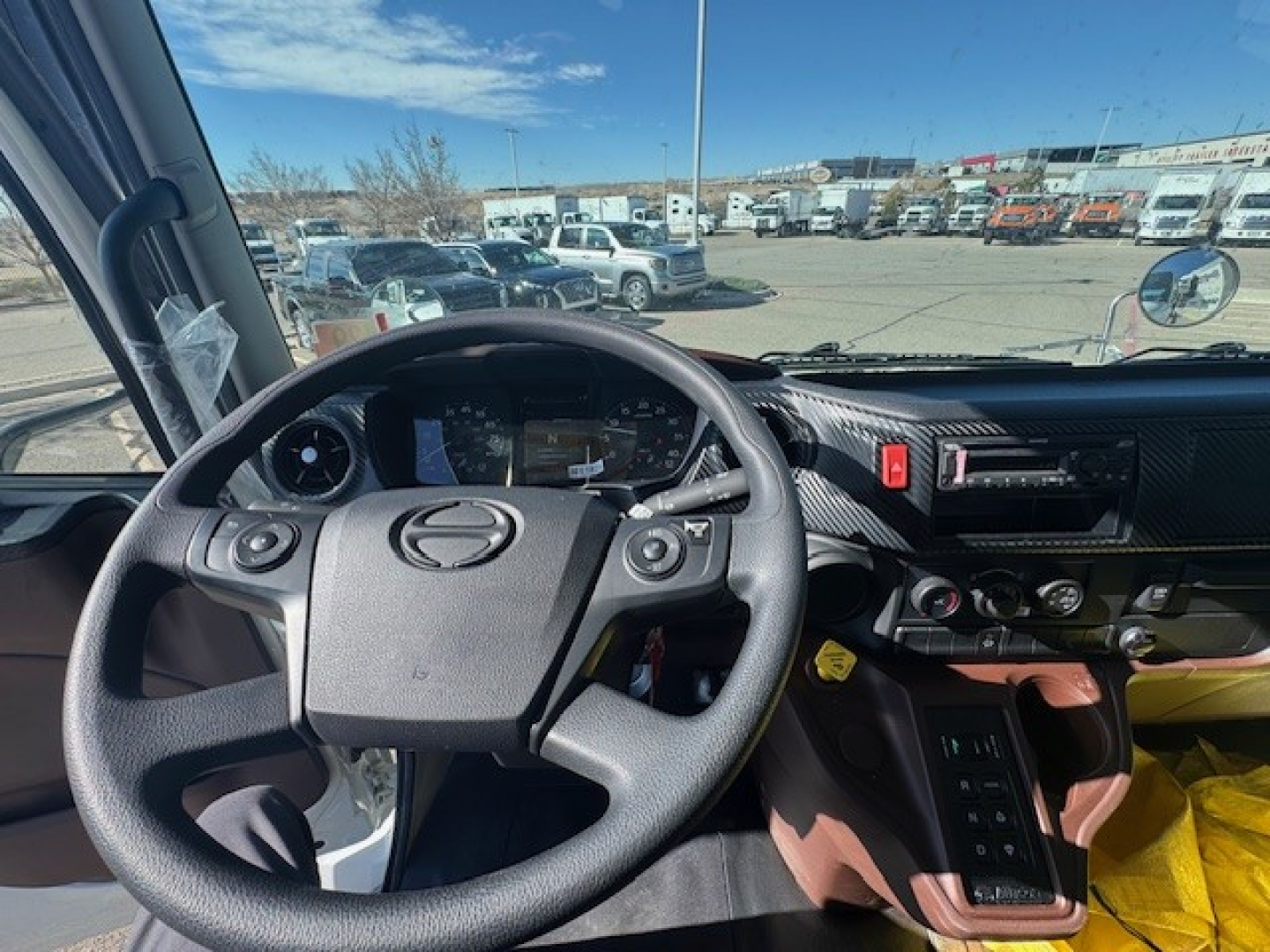 Cab forward view of a 2025 Hino L7 steering wheel and dashboard showing center console controls and gear selector with multiple switches.