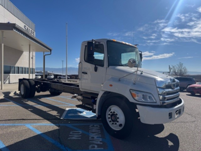 Cab and chassis truck in white, side view showing Hino L7 chassis with bare frame and cab, no cargo bed, dealership lot in background.