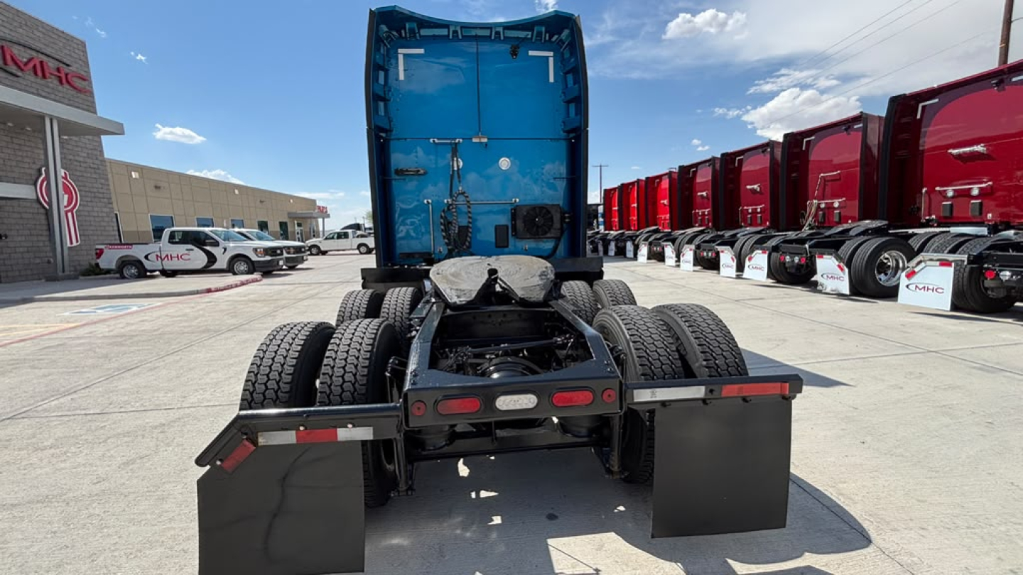 Peterbilt 579 tractor rear view showing fifth wheel, dual rear axles, and mud flaps under a blue sleeper cab with surrounding red trucks.