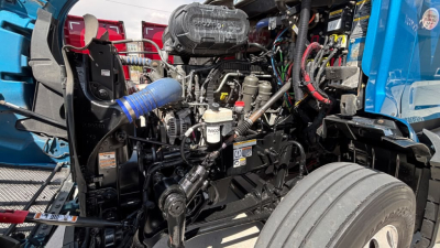 Peterbilt 579 engine bay view showing coolant reservoir and hoses, intake, belts, and front wheel assembly.