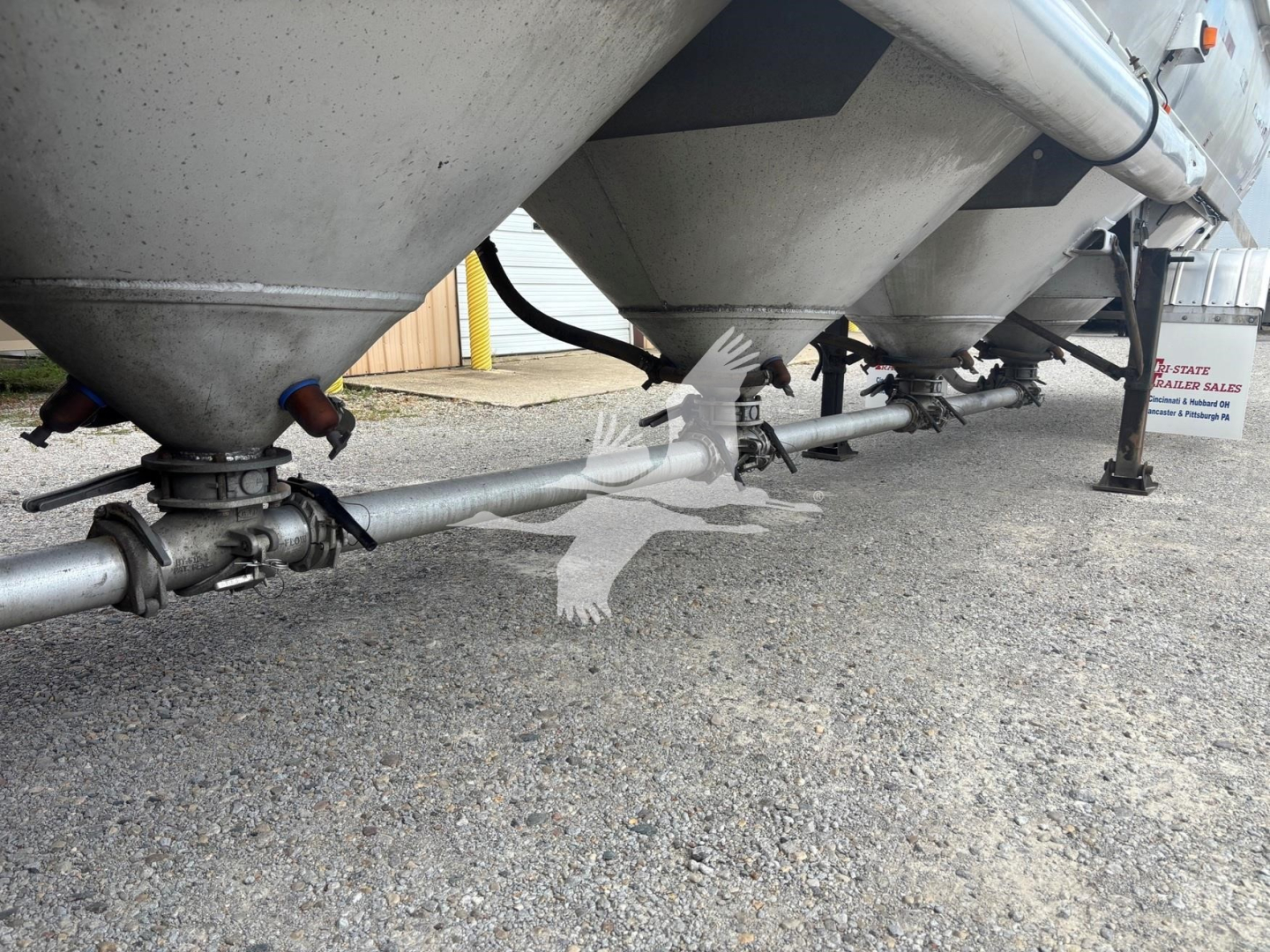 Trailer undercarriage showing multiple conical hoppers and a pneumatic piping run along the bottom with clamps and support legs.