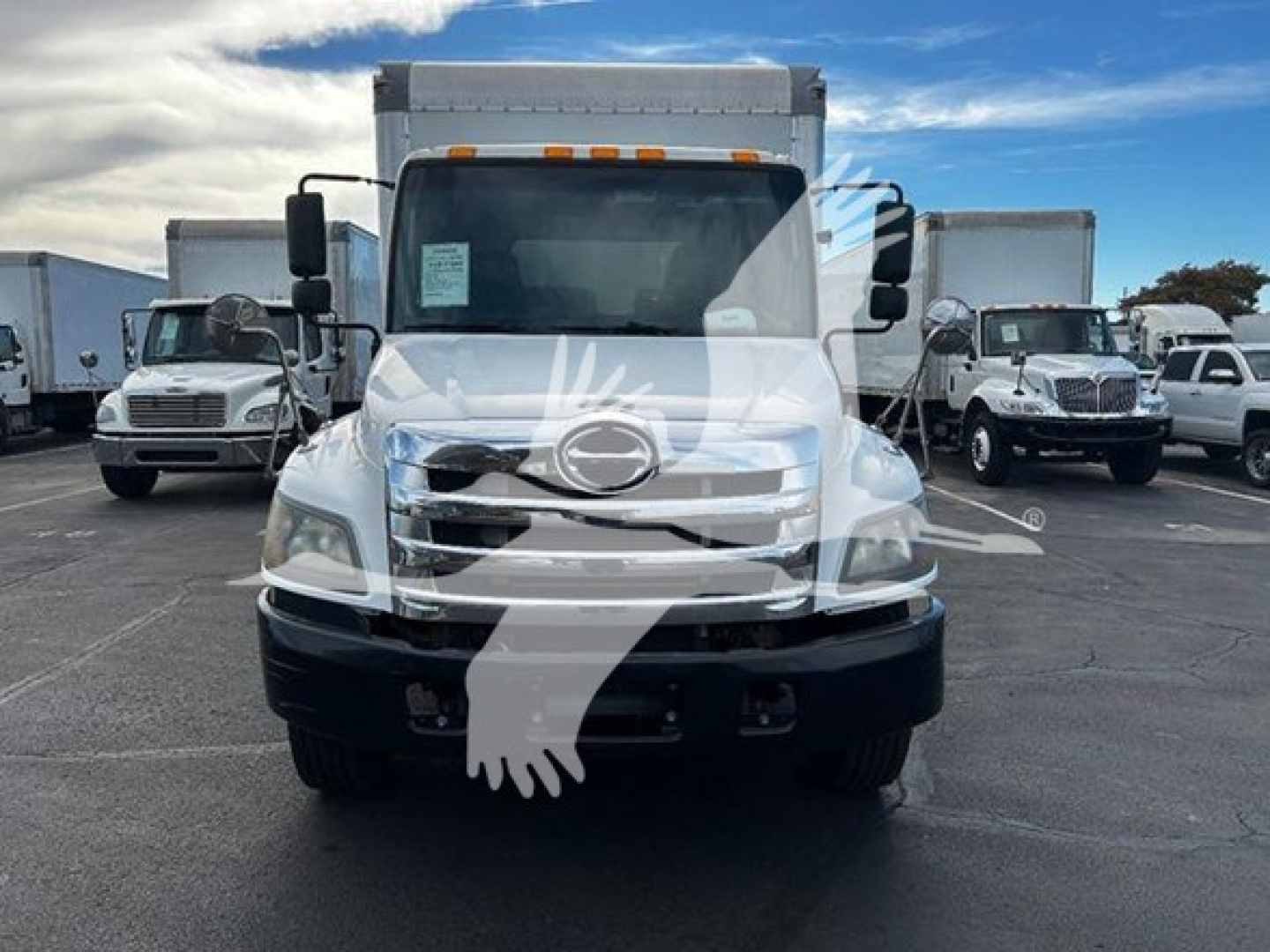 Box truck front view showing Hino logo on grille, headlights, and bumper with elevated cab and windshield; side mirrors visible; other trucks in background.