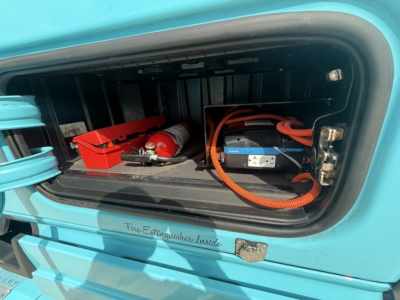 Truck cab storage compartment showing rescue tools and battery pack with orange cable inside a turquoise Freightliner Cascadia 126 engine bay area