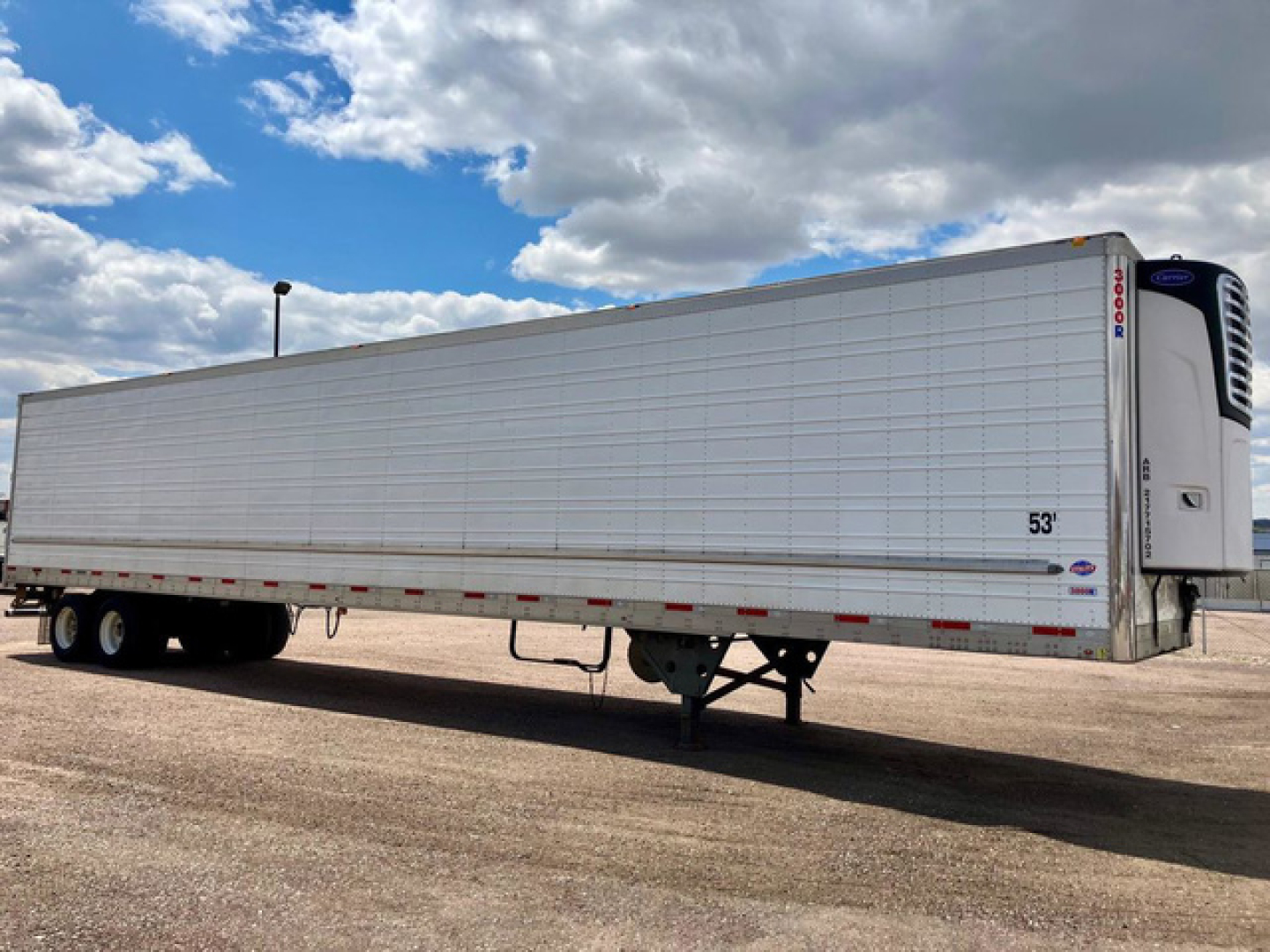 Utility reefer trailer with a white insulated body, 53' length, refrigerated unit at front, tandem axles, parked on dirt under partly cloudy sky.