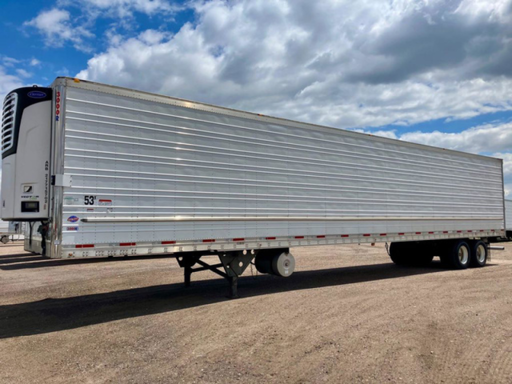Trailer side view of a 2022 Utility reefer trailer with silver aluminum walls, refrigeration unit at front, dual axles, flat terrain and cloudy sky.