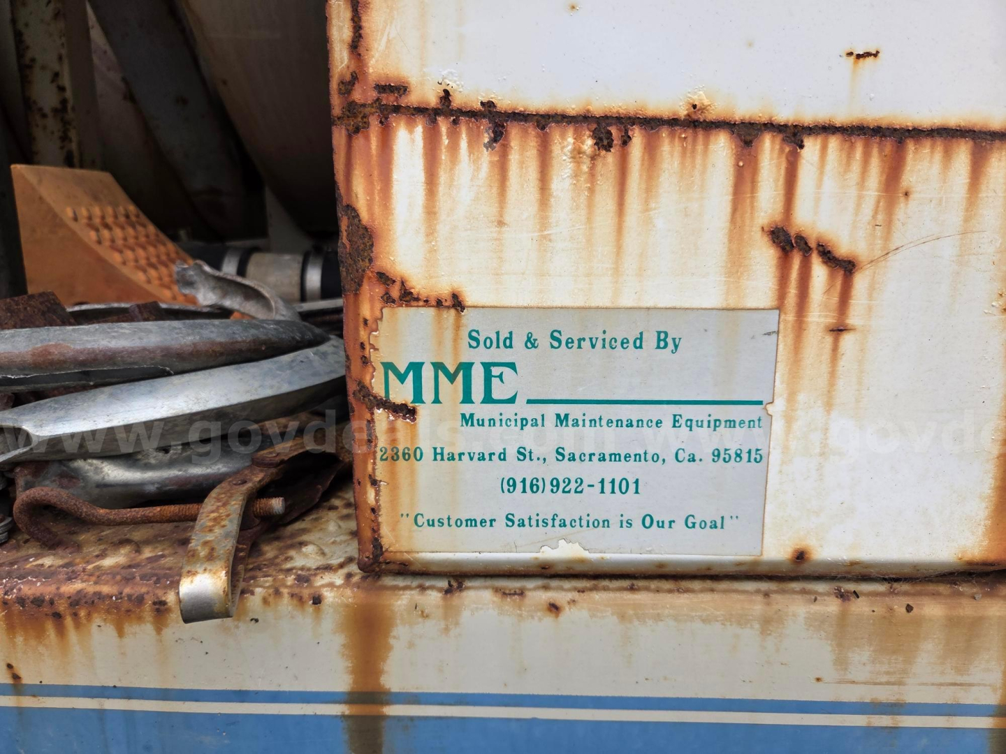 Plaque on a rusted metal equipment showing 'Sold & Serviced By MME Municipal Maintenance Equipment' with address and phone number; rust and grime on surrounding surface.