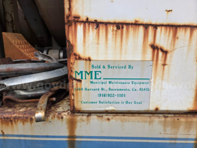 Plaque on a rusted metal equipment showing 'Sold & Serviced By MME Municipal Maintenance Equipment' with address and phone number; rust and grime on surrounding surface.