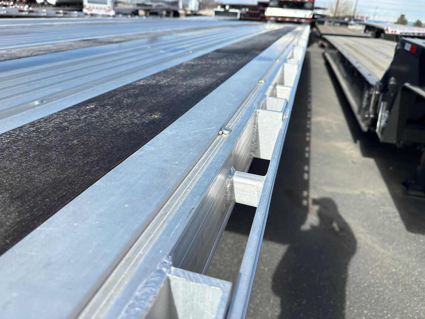 Flatbed trailer bed with metal rails and a black non-slip strip along the surface; corner and side brackets visible; another trailer in the background.