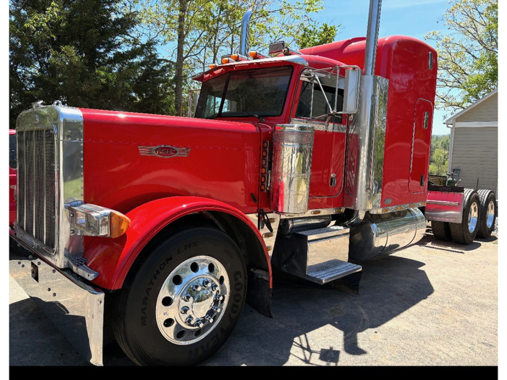 Peterbilt 379 conventional sleeper truck in red, extended hood, chrome exhaust stacks, circular chrome mirrors, front grille, large front tires, daytime outdoor setting.