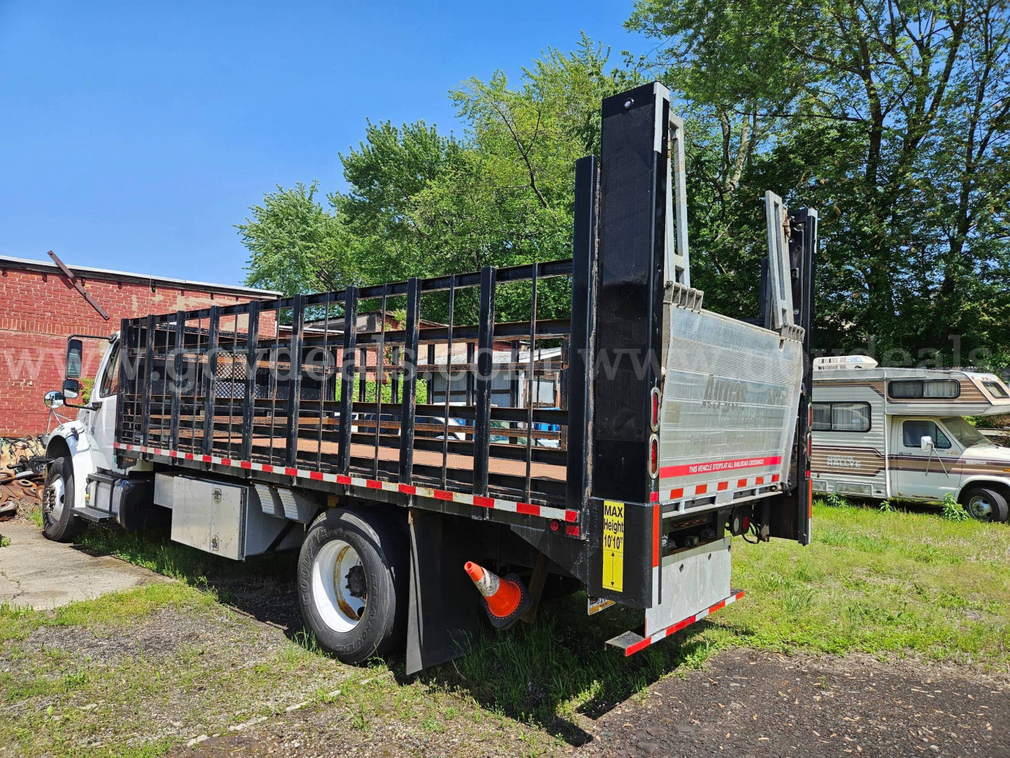 Flatbed truck rear and side view of a 2013 Freightliner M2 106 with high stake bed and hydraulic ramps, showing flatbed, storage boxes, and rear bumper.