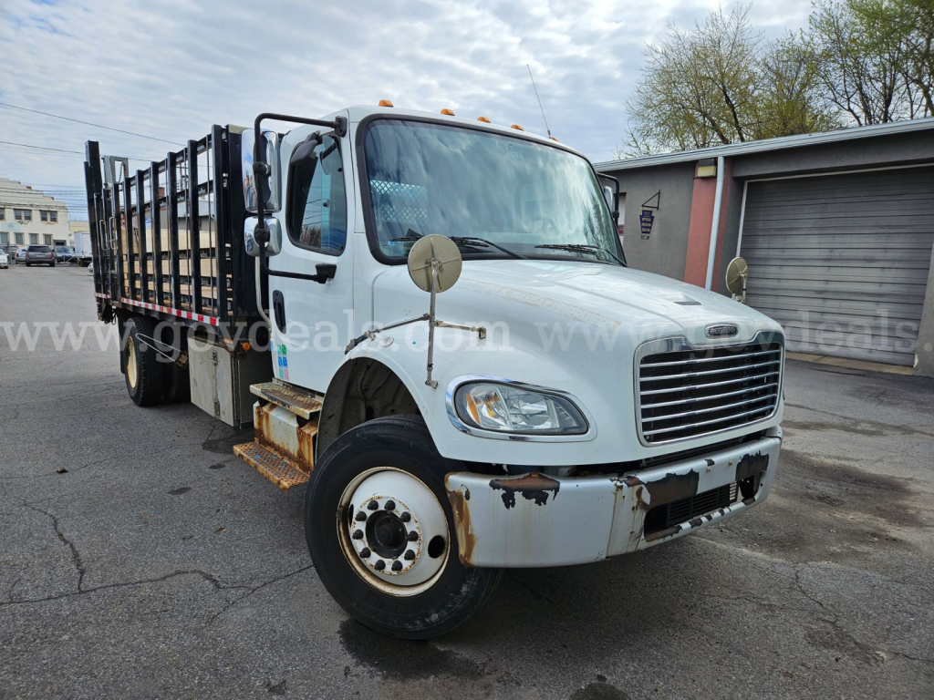 Freightliner M2 106 dump/flatbed truck front-left view with white cab, large grille, side mirrors, and rust on bumper area.