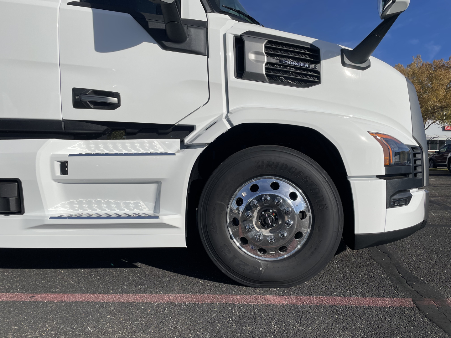 Front side of a white Mack PR64T64 truck showing front wheel and chrome hubcap, cab steps, and part of the bumper.