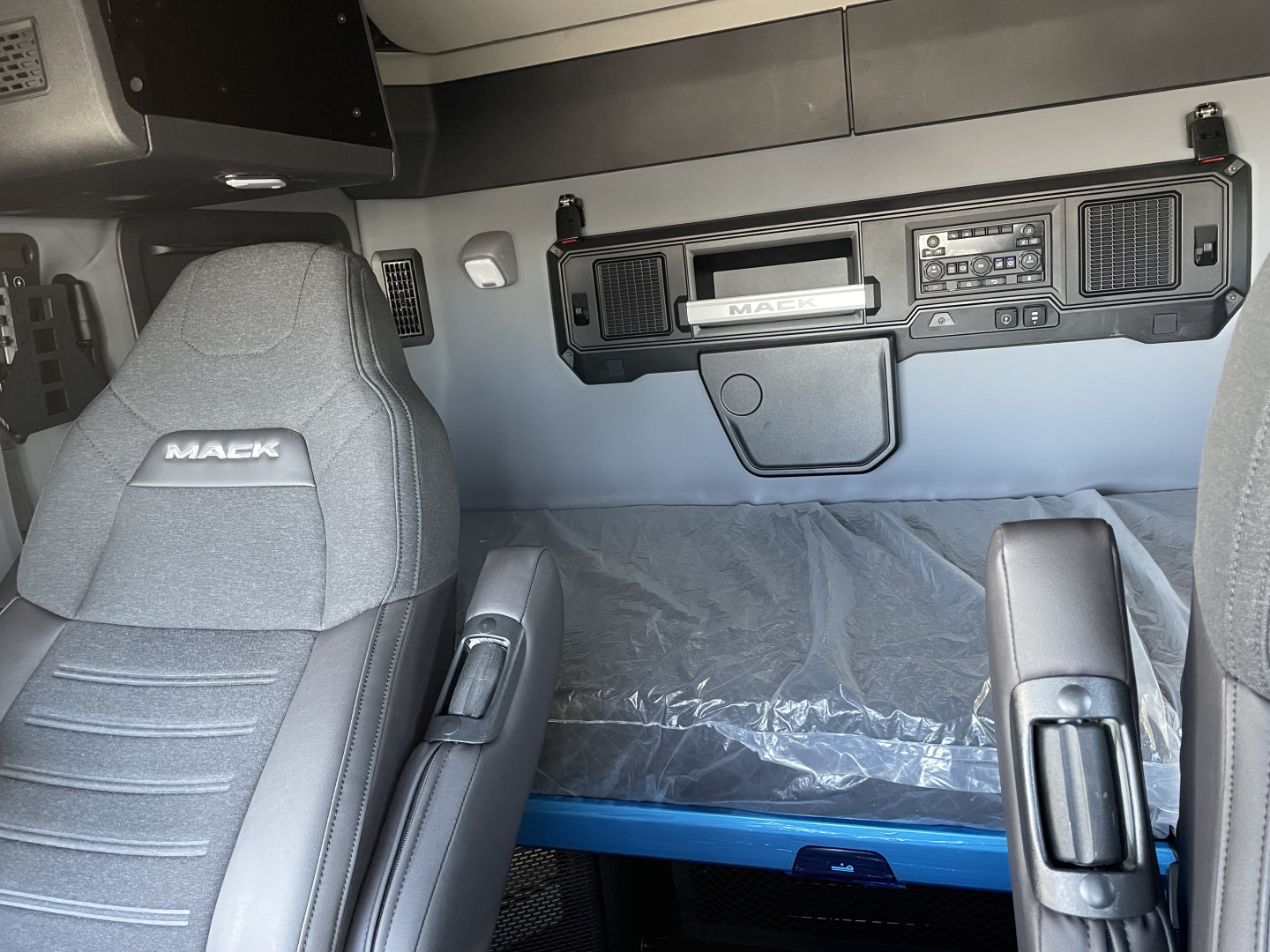 Mack PR64T64 truck interior cab front view showing driver and passenger seats, dashboard with control panel, and sleeper area with plastic cover.