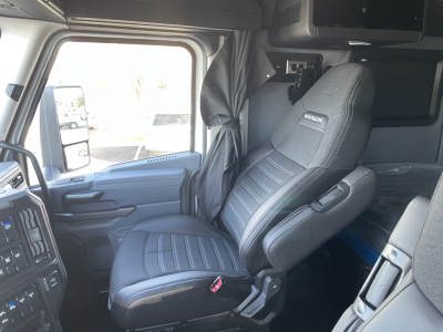 Cab interior of a new 2026 Mack PR64T64 truck showing driver seat with Mack badge and nearby steering wheel, dashboard controls, and sleeping area overhead
