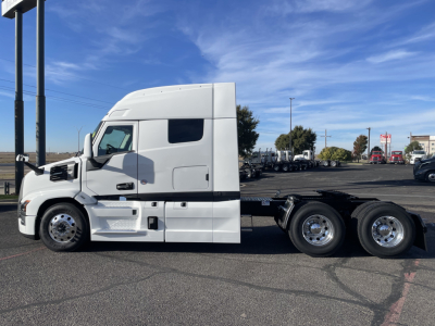truck side view with a white 2026 Mack PR64T64 conventional sleeper cab on a trailer chassis, visible wheels, and dealership lot in background.