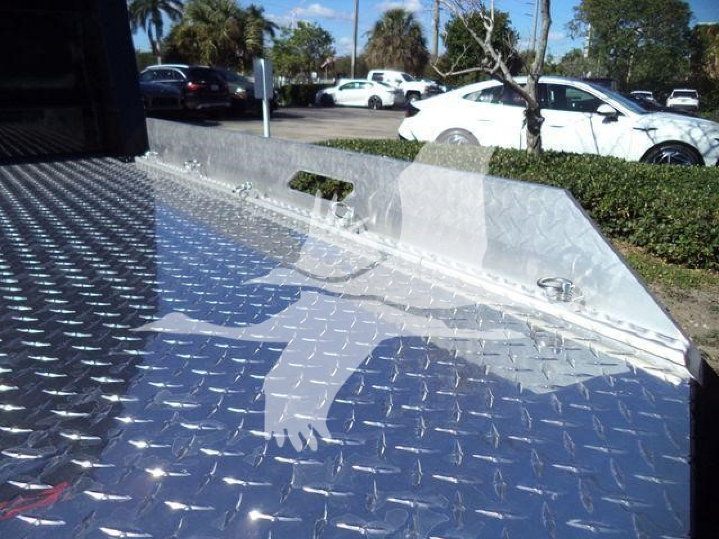 Flatbed truck bed with diamond plate pattern; front side shows raised aluminum compartment and raised winch area. Background includes parked cars and trees.