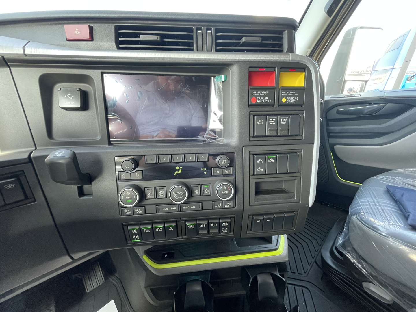 Truck dash with multiple control panels showing HVAC knobs, infotainment screen, various switches, and storage area. Visible text includes warning labels and control symbols on center console.