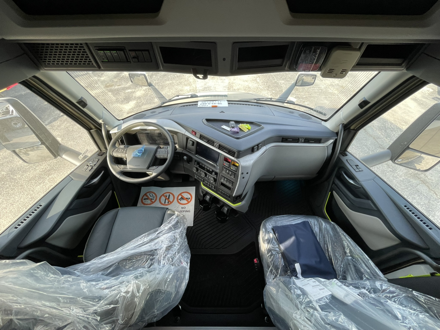 Volvo VNL64T860 truck cab interior showing steering wheel, dashboard controls, and plastic-wrapped seats, front view from driver seat.