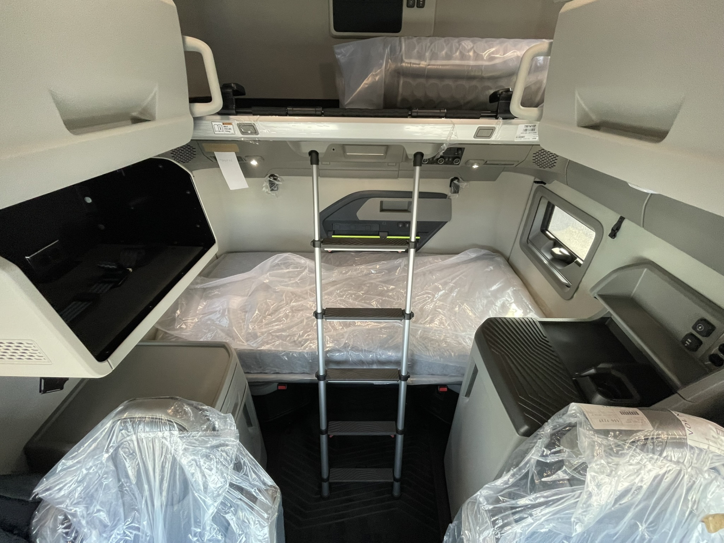 Interior of a new Volvo VNL sleeper cab showing bunk bed with ladder, overhead storage, and plastic-covered seating; control panels and window visible.