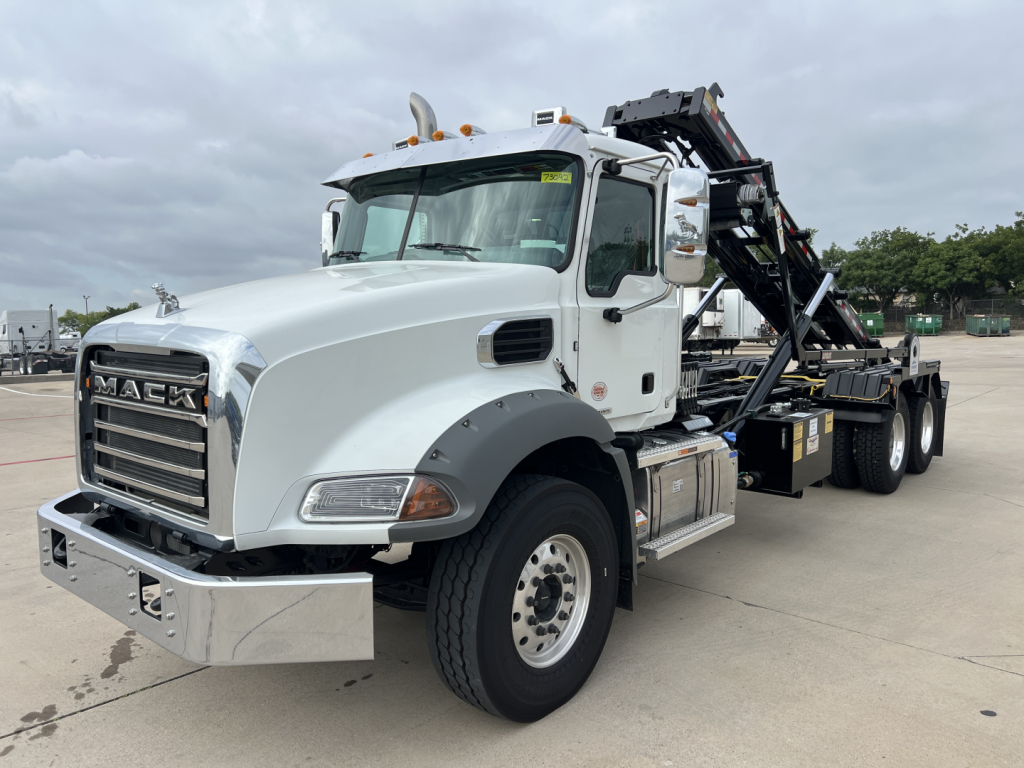 Mack GR64B roll-off truck in white with a hydraulic lifting bed and large front bumper, daytime setting.