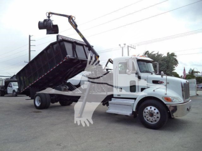 Peterbilt 337 truck with a hydraulic grapple lifting a black dump bed, cab white, side view showing crane arm and hook in operation.