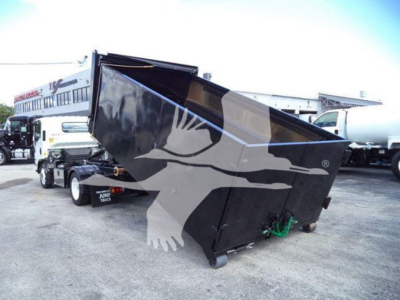 Isuzu NPR-HD truck in a yard with a large black dump body/box attached, tilt/open top, side view, other trucks in background.
