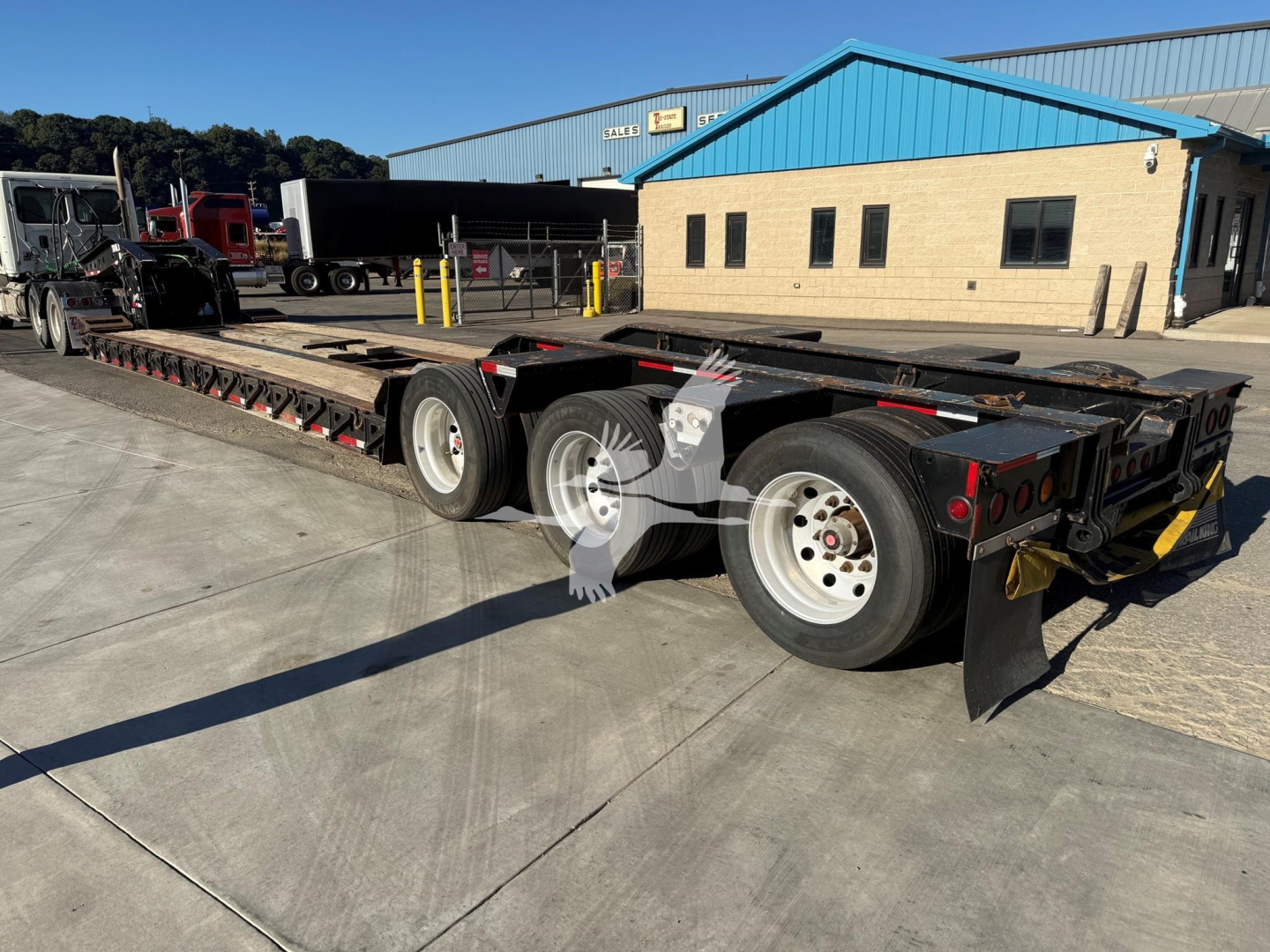 Trailer with lowboy flatbed showing multiple axles and tires, rear ramps, and safety reflectors. No legible labels visible in close-up text.