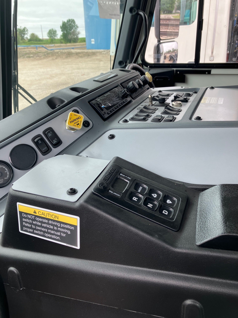 Cockpit of a 2025 Mack LR64 cab showing control panel with switches and digital cluster, warning label, and steering area.