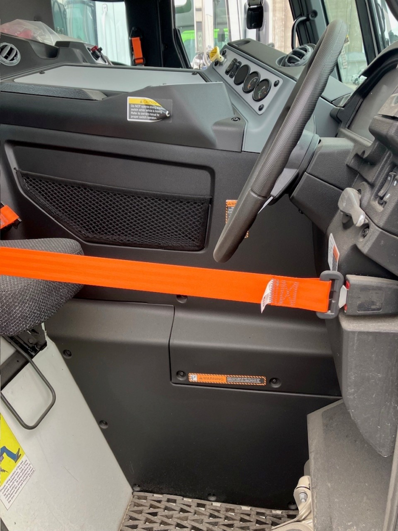 Truck cab interior showing steering wheel and dashboard of a new 2025 Mack LR64, with orange seat belt across the doorway and control cluster visible.