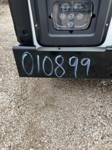 Front bumper area of a 2025 Mack LR64 cab and chassis showing the rear tail/step area with the stock number 010899 written on the bumper and a light housing.