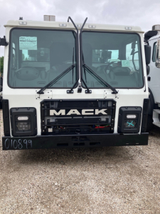 Mack LR64 cab and chassis front showing large grille with 'MACK' badge, dual windshield wipers, and white cab; VIN sticker on left window and stock number on windshield.