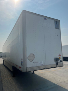 Rear view of a white utility van trailer showing a small door latch and safety placards on the back, smooth metal exterior panels and reflective decals.