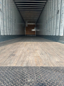 Truck trailer interior showing empty cargo area with wooden floor and metal side walls; open loading door at far end.
