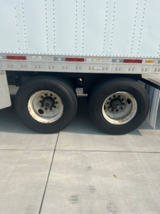 Dual rear wheels of a utility trailer with a white enclosed cargo body, showing wheel rims and tires; side of trailer with riveted panels and reflective markers.