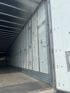 Inside of an empty trailer van with open rear doors, showing metal interior walls and wooden floor.