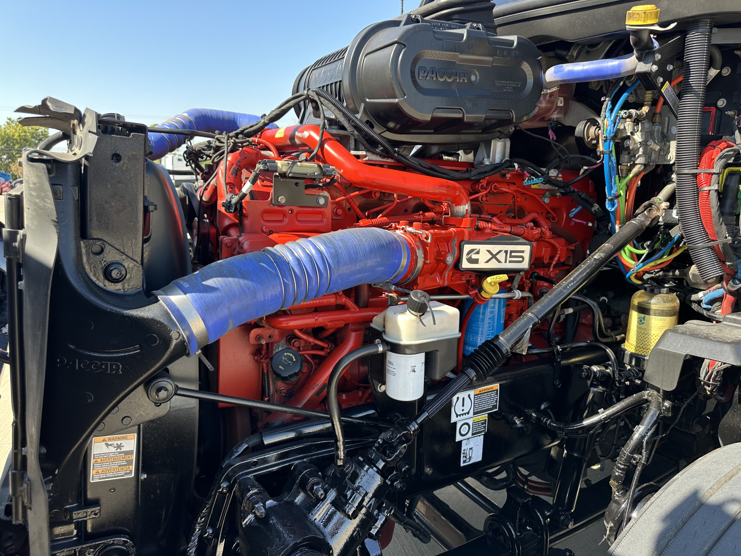 Kenworth T680 engine bay with red engine, blue intake hose, and various hoses and components visible.