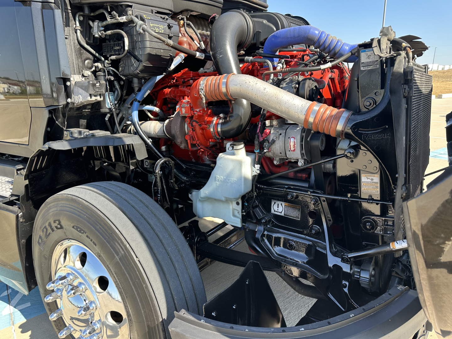 Kenworth T680 engine bay showing red V8 engine, intake hoses, radiator, and front wheel with chrome hubcap.