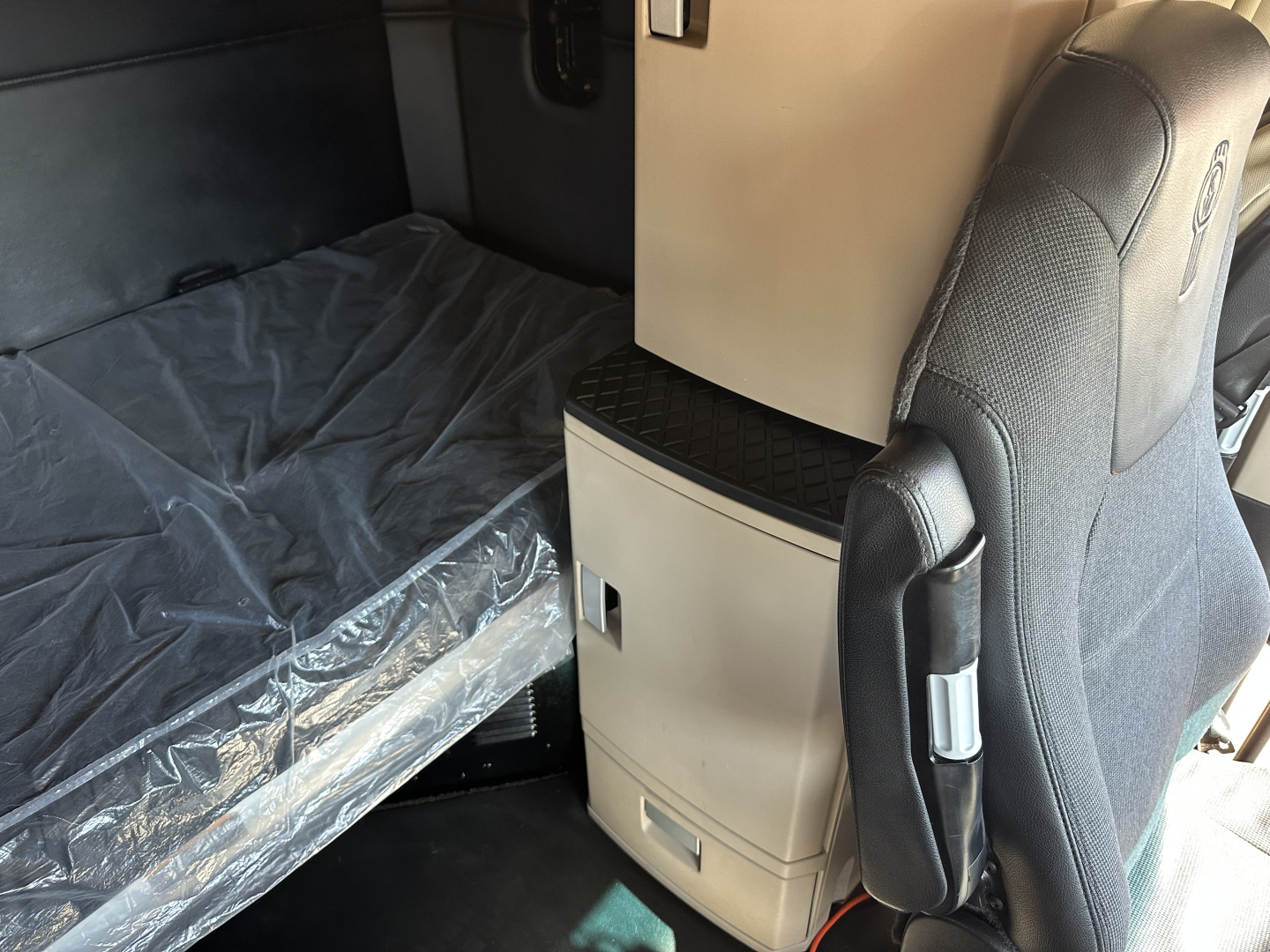 Kenworth T680 interior cabin area with a plastic-covered sleeper bed and center console; seats and storage cabinet visible.