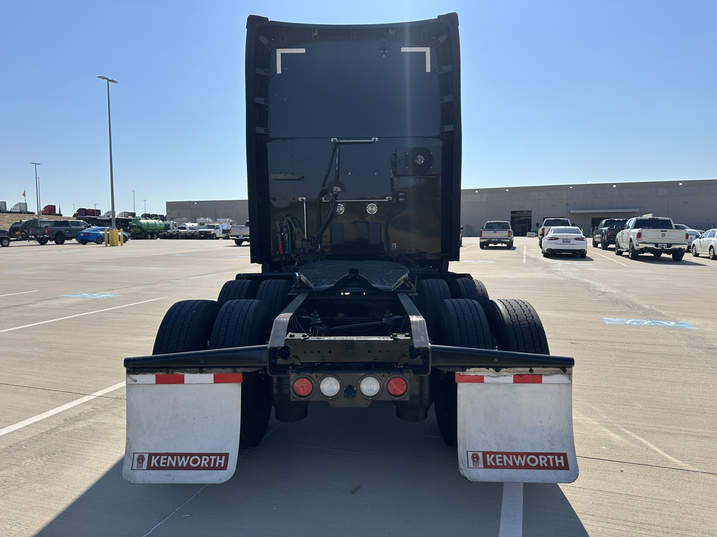Rear view of a 2022 Kenworth T680 truck, showing the back of the cab and dual rear wheels. Mudflaps labeled 'KENWORTH' on both sides, with red and white reflective strips on the rear bumper.