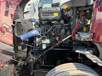 Kenworth T680 engine bay showing Denso air intake and various hoses with yellow warning label, exposed frame and front wheel, hood open.