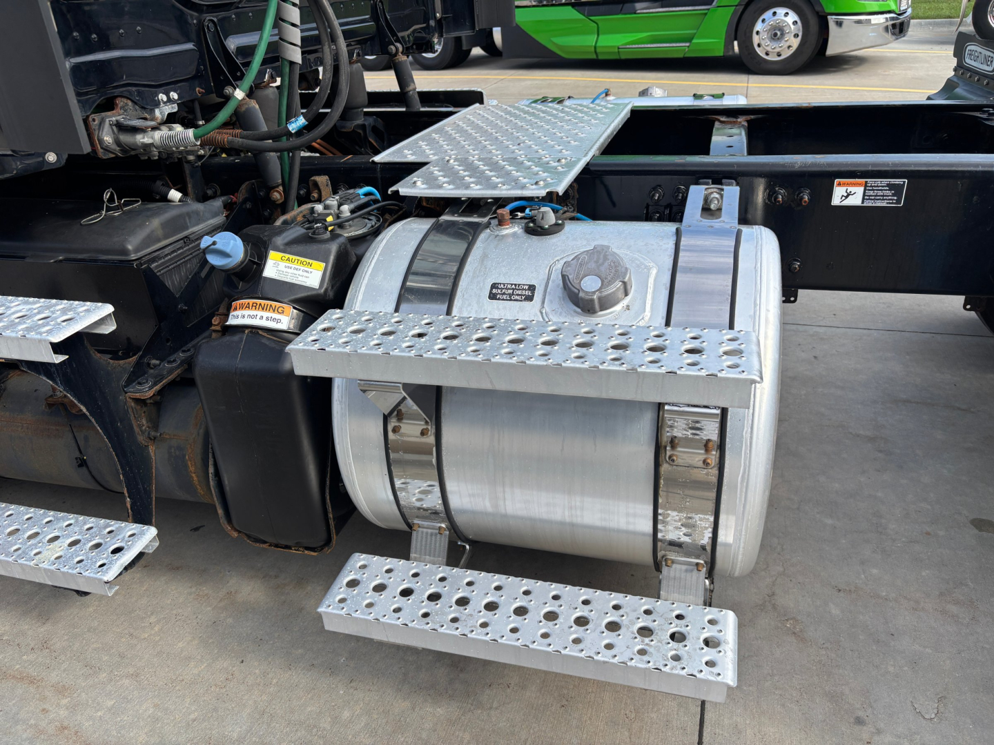Fuel tank and access steps on a Freightliner truck undercarriage with metal perforated steps and cab area visible.
