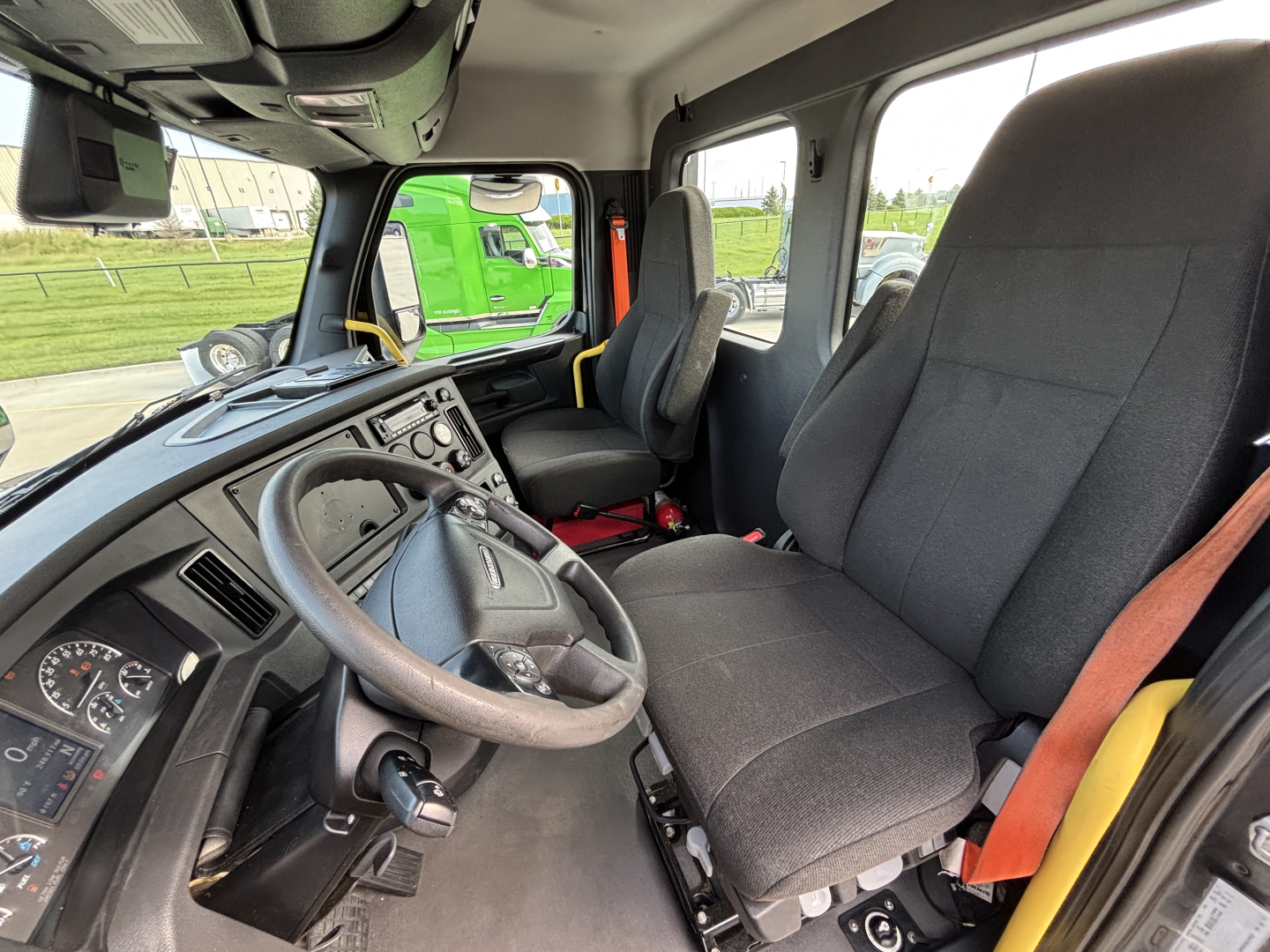 Freightliner cab interior showing steering wheel, driver seat, and dashboard with gauges; front passenger seat and side window visible.