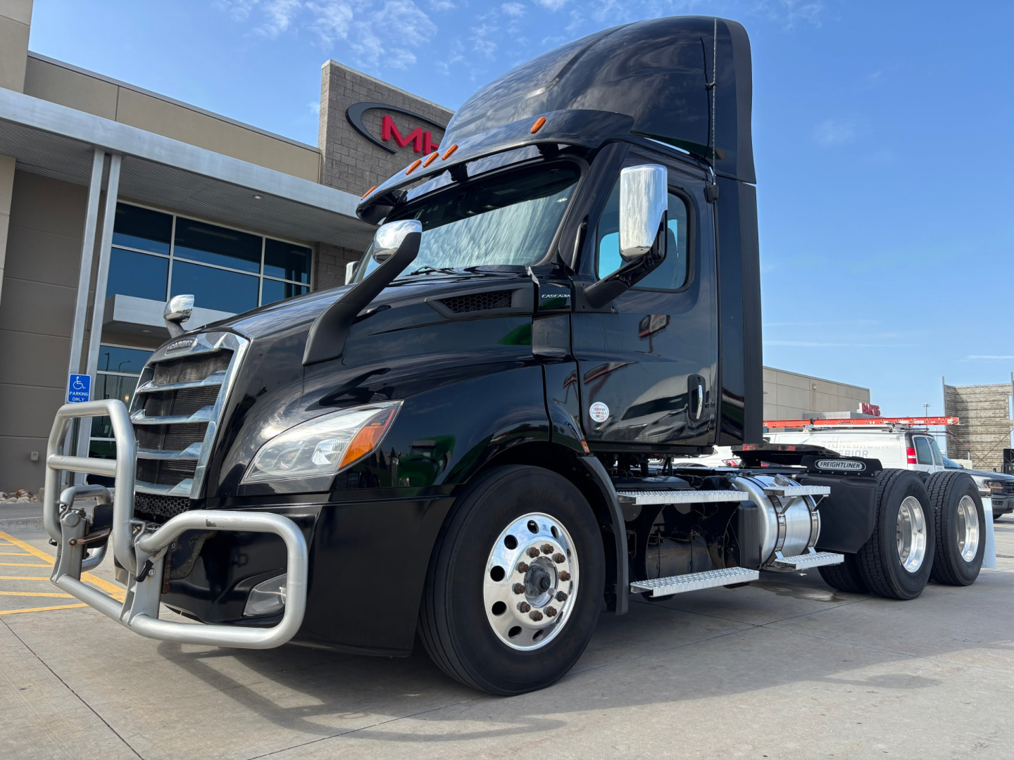 Freightliner conventional daycab tractor in black, front grille guard, side mirrors, dual rear axles, fuel tanks, and aggressive hood; showing cab and chassis in a dealership lot.