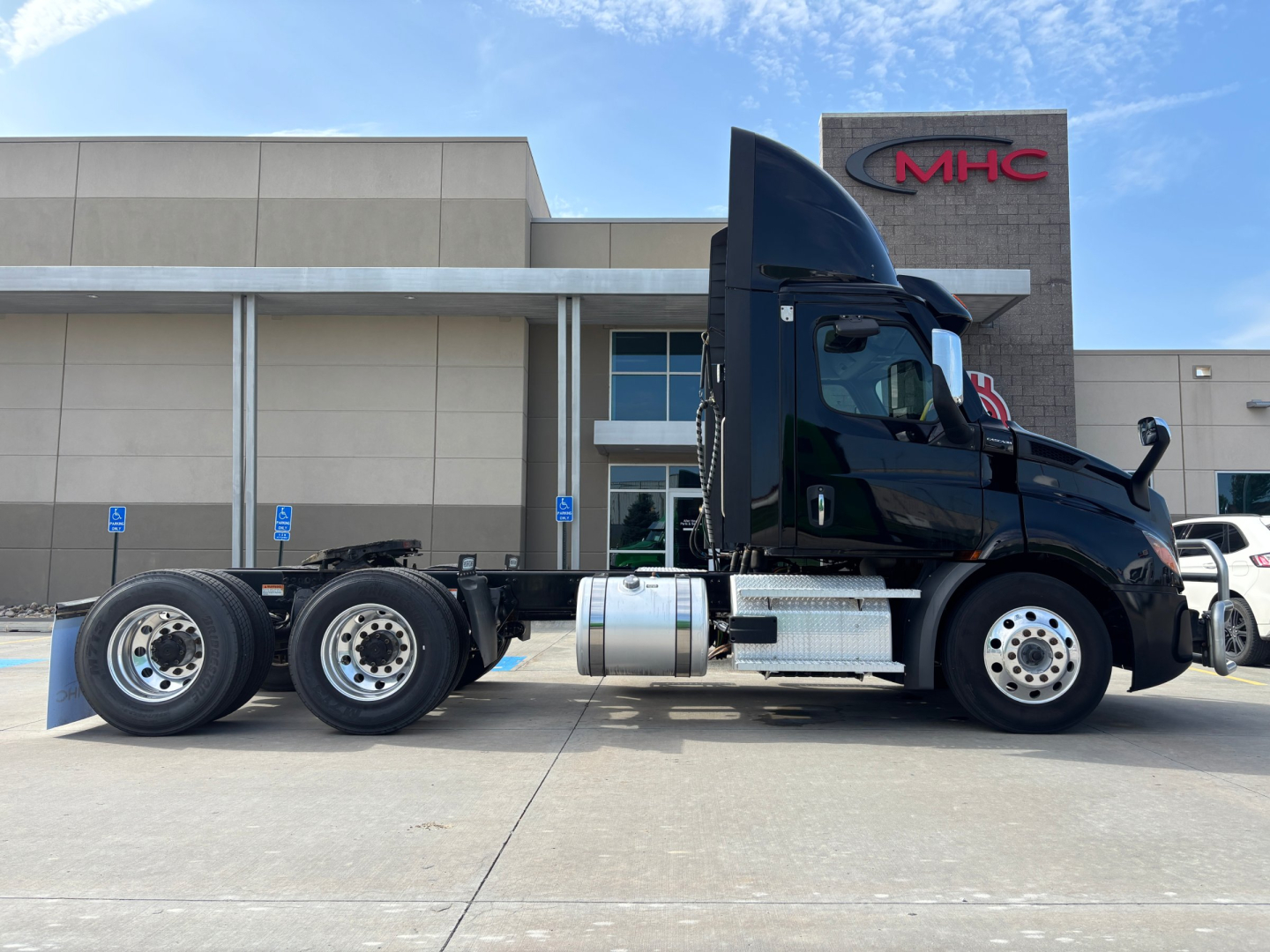 Truck side profile of a 2023 Freightliner conventional daycab with dual rear axles and turquoise tank, parked in front of a building with MHC sign.